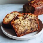 Two moist slices of chocolate chip banana bread recipe on a white plate, with the loaf visible in the background.