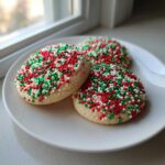 Three round christmas sprinkle sugar cookies covered in red, green, and white nonpareils, served on a white plate.