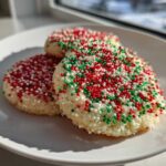 Three round christmas sprinkle sugar cookies covered in red, green, and white nonpareils on a white plate.