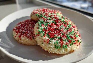 Three round christmas sprinkle sugar cookies covered in red, green, and white nonpareils on a white plate.