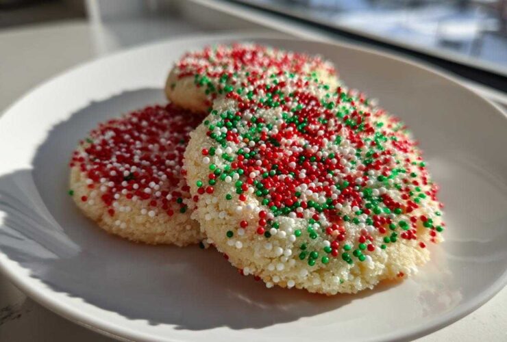 Three round christmas sprinkle sugar cookies covered in red, green, and white nonpareils on a white plate.