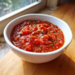 A close-up of chunky, vibrant red pizza sauce seasoned with herbs and pepper flakes in a white bowl.