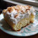 A moist slice of coconut poke cake topped with whipped cream and toasted coconut flakes.