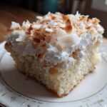 A moist slice of coconut poke cake topped with whipped cream and toasted coconut flakes on a decorative plate.