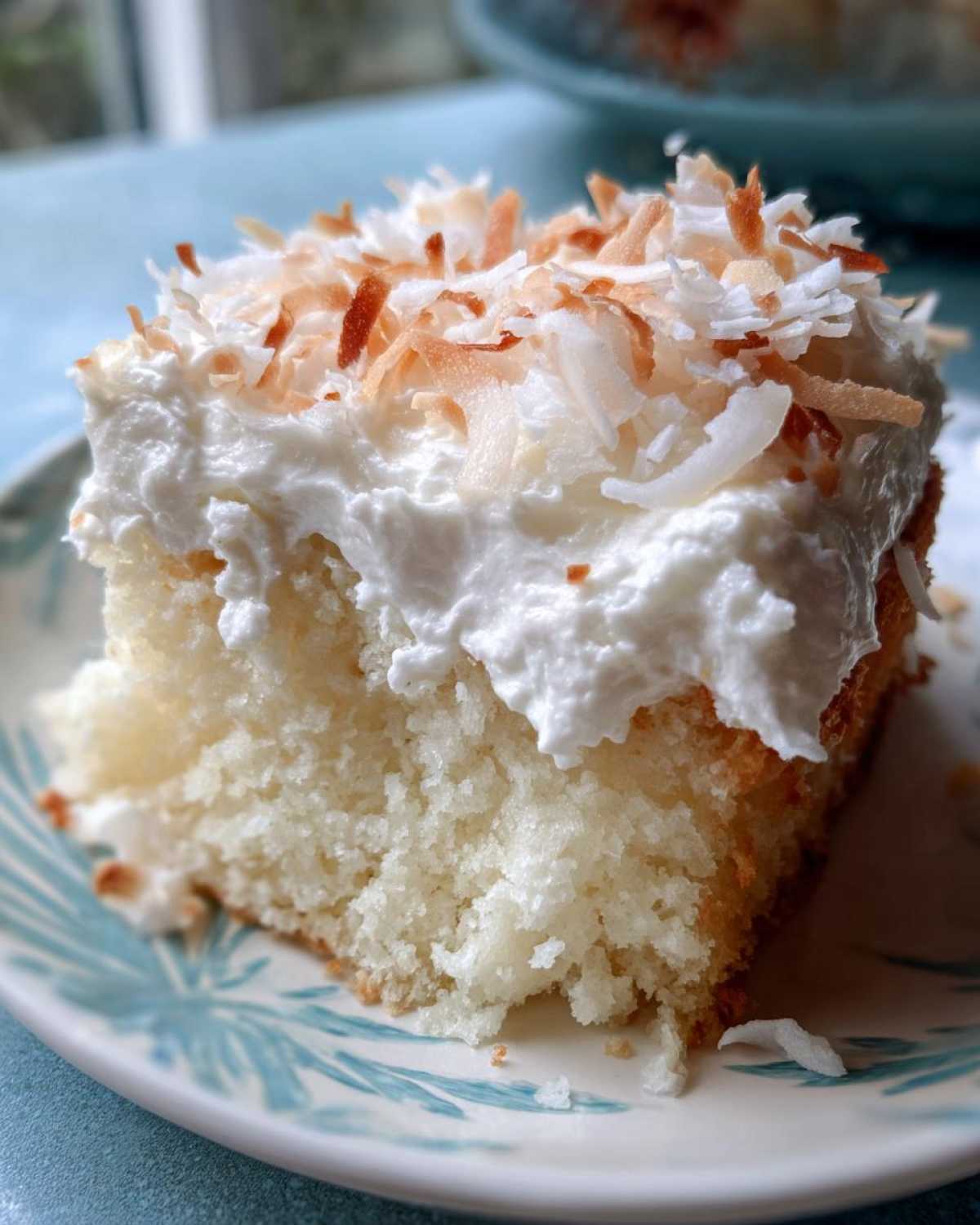 Close-up of a moist slice of coconut poke cake topped with whipped cream and toasted coconut flakes.
