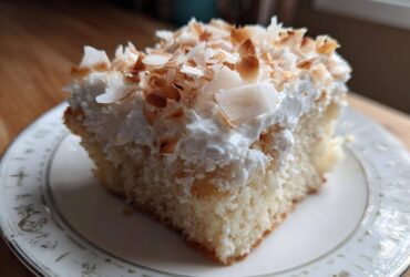 A moist slice of coconut poke cake topped with whipped cream and toasted coconut flakes on a decorative plate.
