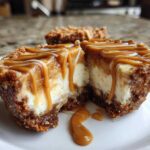 A close-up of a cookie butter cheesecake cup cut in half, showing creamy filling and a thick cookie crust, drizzled heavily with caramel sauce.