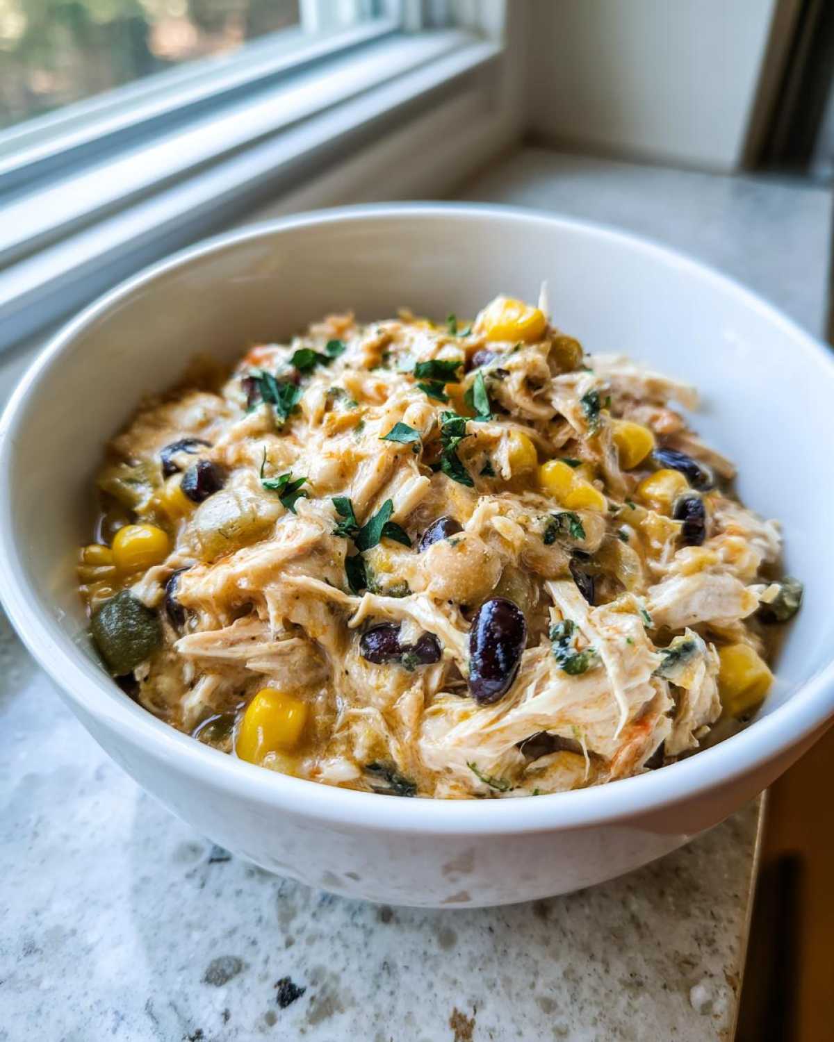 A close-up of a bowl filled with creamy, shredded cream cheese chicken chili, corn, and black beans, garnished with parsley.