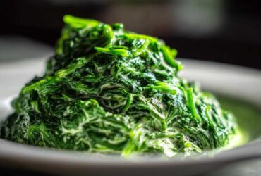 A mound of vibrant green, rich creamed spinach recipe served on a white plate, showing the creamy sauce coating the leaves.