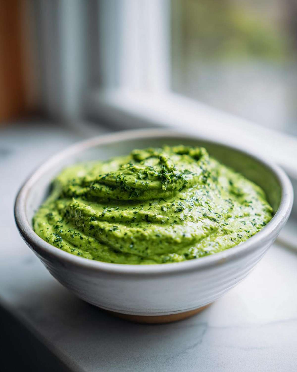 A close-up of thick, vibrant green creamy avocado sauce topped with chopped herbs in a rustic white bowl.