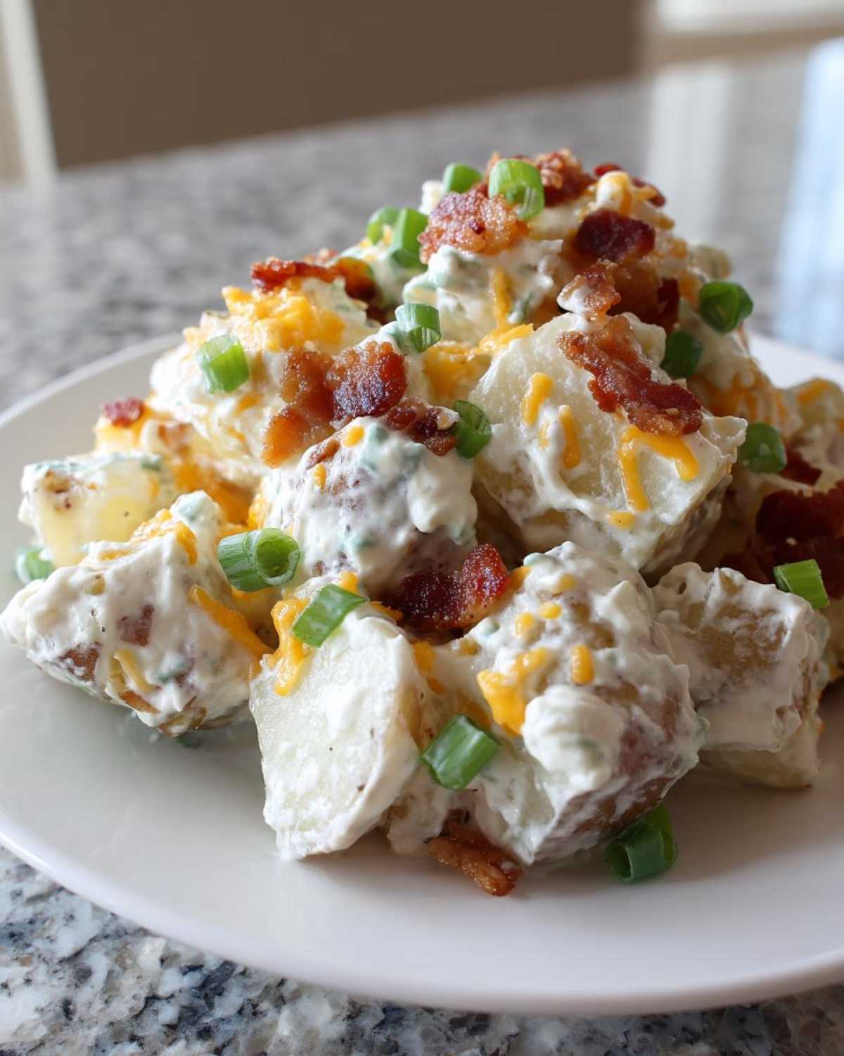 Close-up of a mound of creamy baked potato salad topped with bacon bits, shredded cheddar cheese, and green onions.