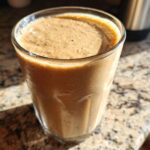 Close-up of a creamy brown banana smoothie in a clear glass sitting on a granite countertop.
