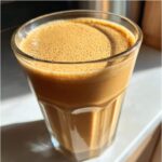 Close-up of a creamy, light brown banana smoothie with a frothy top in a clear glass.