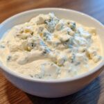 Close-up of a white bowl filled with creamy blue cheese dip, topped generously with large crumbles of blue cheese.