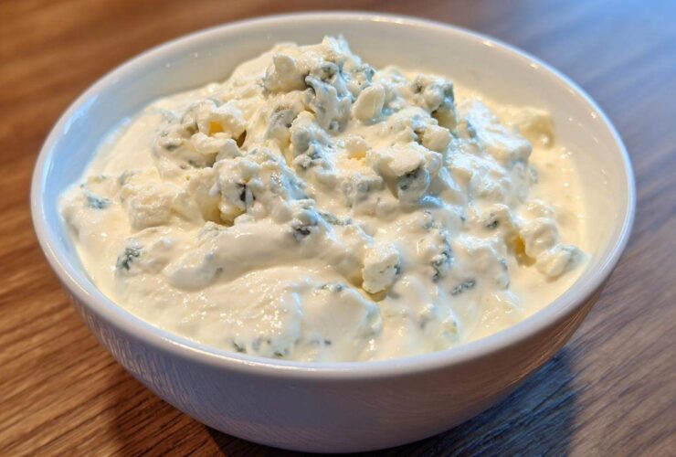 Close-up of a white bowl filled with creamy blue cheese dip, topped generously with large crumbles of blue cheese.