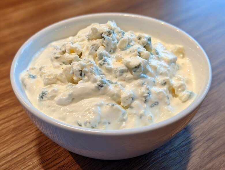 Close-up of a white bowl filled with creamy blue cheese dip, topped generously with large crumbles of blue cheese.