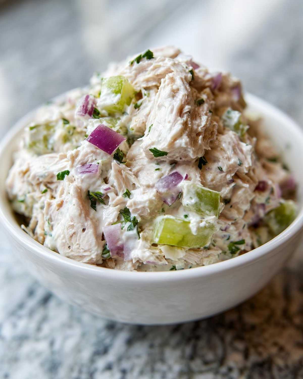 Close-up of a creamy healthy tuna salad mixture with visible chunks of celery, red onion, and herbs in a white bowl.