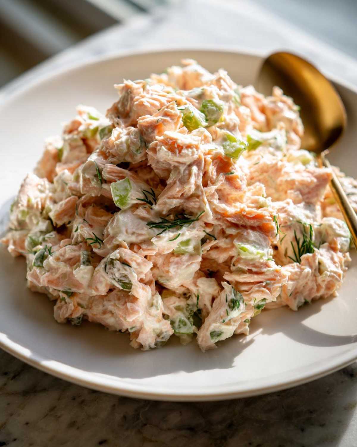 A mound of creamy salmon salad mixed with celery and dill served on a white plate.