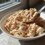 A spoonful of creamy salmon salad, mixed with celery and dill, being lifted from a white bowl.