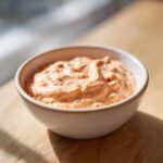 A close-up of creamy, orange-hued smash burger sauce speckled with herbs in a small white bowl.