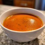 Close-up of a steaming bowl of rich, orange-red tomato bisque, garnished with pepper.