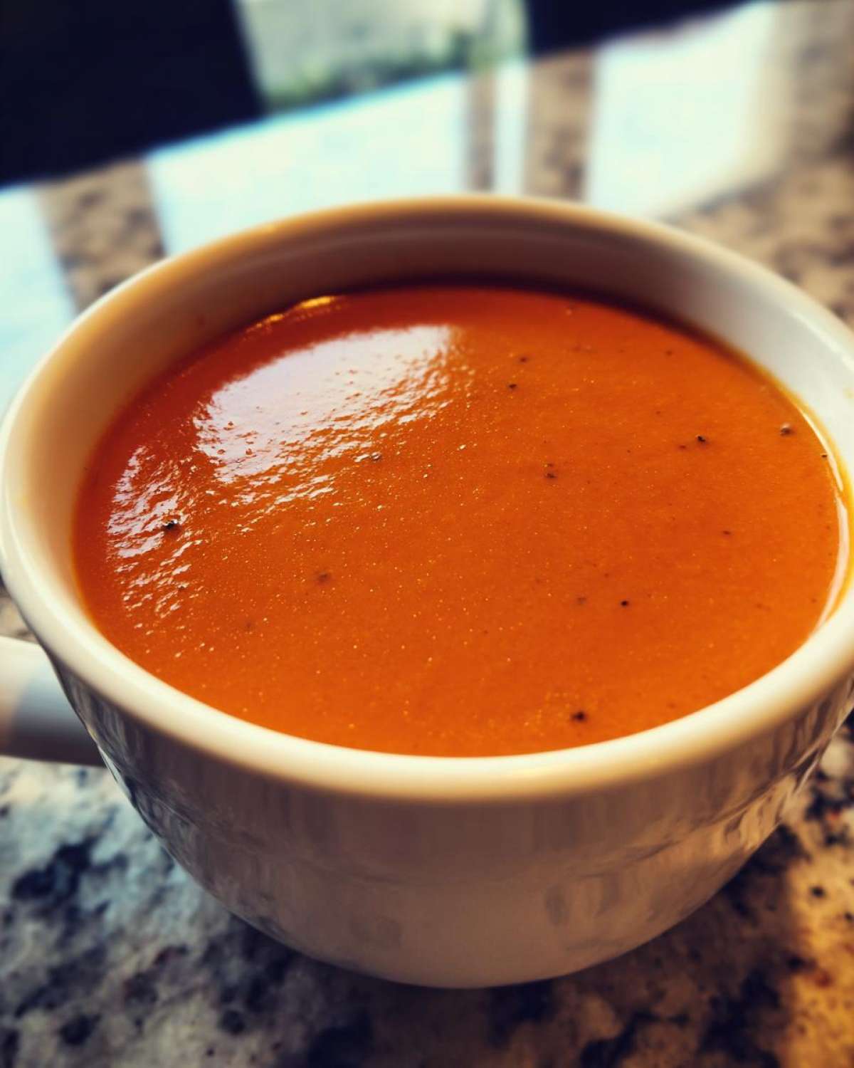 Close-up of rich, orange-red tomato bisque soup served hot in a white mug on a granite countertop.