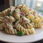 A close-up of creamy tuna pasta salad made with fusilli, peas, and red onion on a white plate.