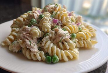 A close-up of creamy tuna pasta salad made with fusilli, peas, and red onion on a white plate.