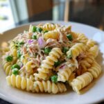 Close-up of creamy Tuna pasta salad featuring rotini pasta, shredded tuna, green peas, and diced red onion on a white plate.