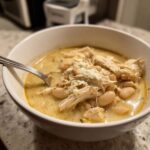A close-up of a white bowl filled with creamy white chicken chili, featuring shredded chicken, white beans, and a sprinkle of cheese.