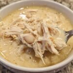 A spoonful of creamy white chicken chili showing shredded chicken and white beans being lifted from a white bowl.