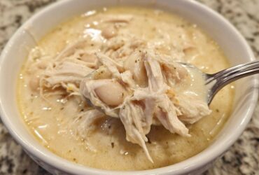 A spoonful of creamy white chicken chili showing shredded chicken and white beans being lifted from a white bowl.