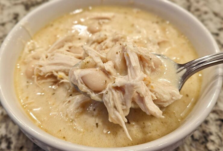 A spoonful of creamy white chicken chili showing shredded chicken and white beans being lifted from a white bowl.