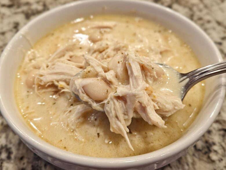 A spoonful of creamy white chicken chili showing shredded chicken and white beans being lifted from a white bowl.