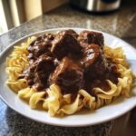 Close-up of tender crockpot beef tips smothered in rich brown gravy served over egg noodles on a white plate.