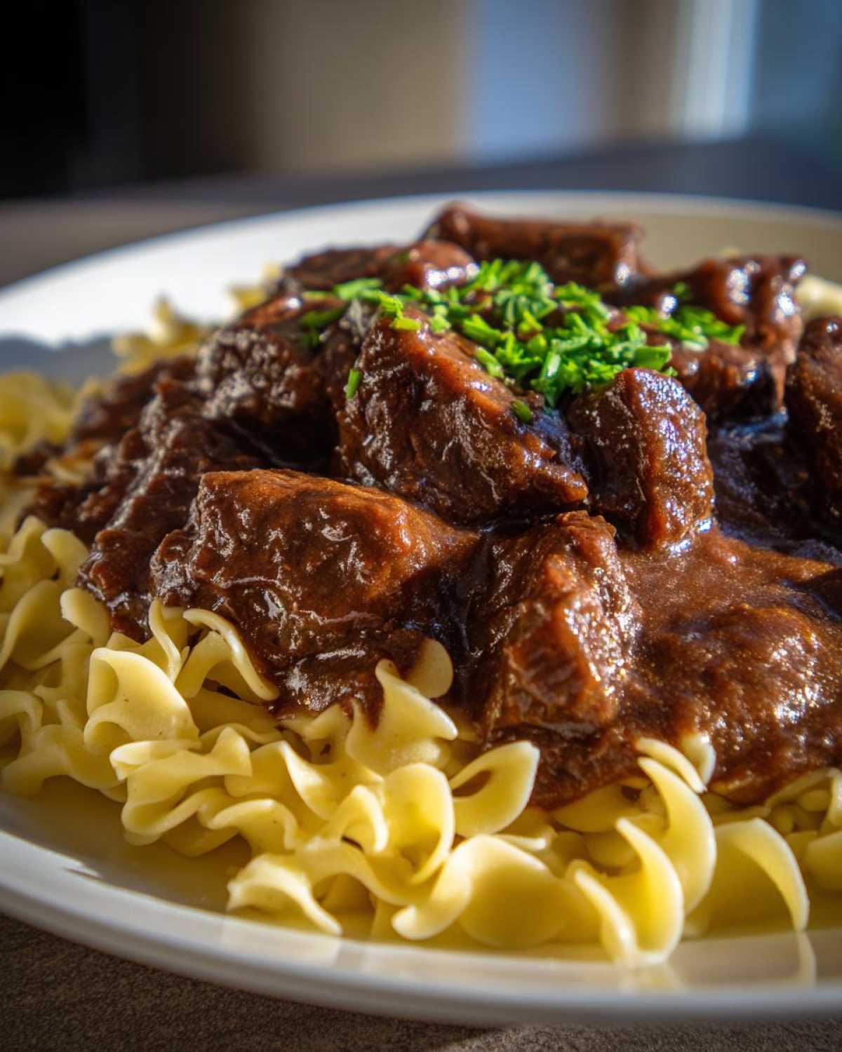 Close-up of rich, dark crockpot beef tips smothered in gravy served over wide egg noodles and topped with fresh chives.