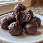 A pile of rich, chocolate-covered pecan pie balls on a white plate, one is broken open showing the filling.