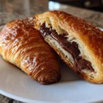 Close-up of a flaky, golden brown chocolate croissant cut in half, revealing rich melted chocolate filling.