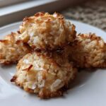 Four golden-brown almond joy cookies stacked slightly on a white plate, showing toasted coconut texture.