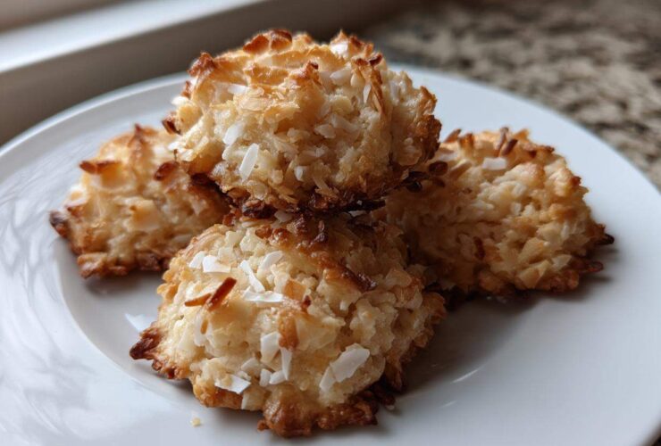 Four golden-brown almond joy cookies stacked slightly on a white plate, showing toasted coconut texture.