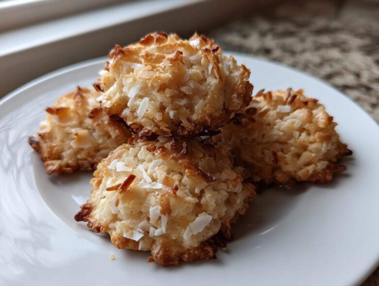 Four golden-brown almond joy cookies stacked slightly on a white plate, showing toasted coconut texture.