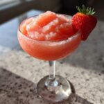 A vibrant, frozen strawberry daiquiri served in a coupe glass, garnished with a fresh strawberry.