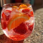 Close-up of a glass filled with bubbly champagne punch, ice, sliced strawberries, and orange wedges.