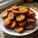 A mound of perfectly golden brown, crispy fried okra slices piled high on a white plate.