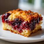Close-up of a gooey square slice of cherry dump cake with a golden, crumbly topping on a white plate.