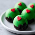 Close-up of several bright green Grinch Oreo Balls dipped in chocolate crumbs and topped with a small red heart.