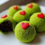 A plate featuring several bright green Grinch Oreo Balls, one of which is broken open showing the dark chocolate interior, topped with small red hearts.