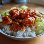 A bowl featuring saucy, glazed harissa chicken pieces served over white rice and surrounded by crisp lettuce.