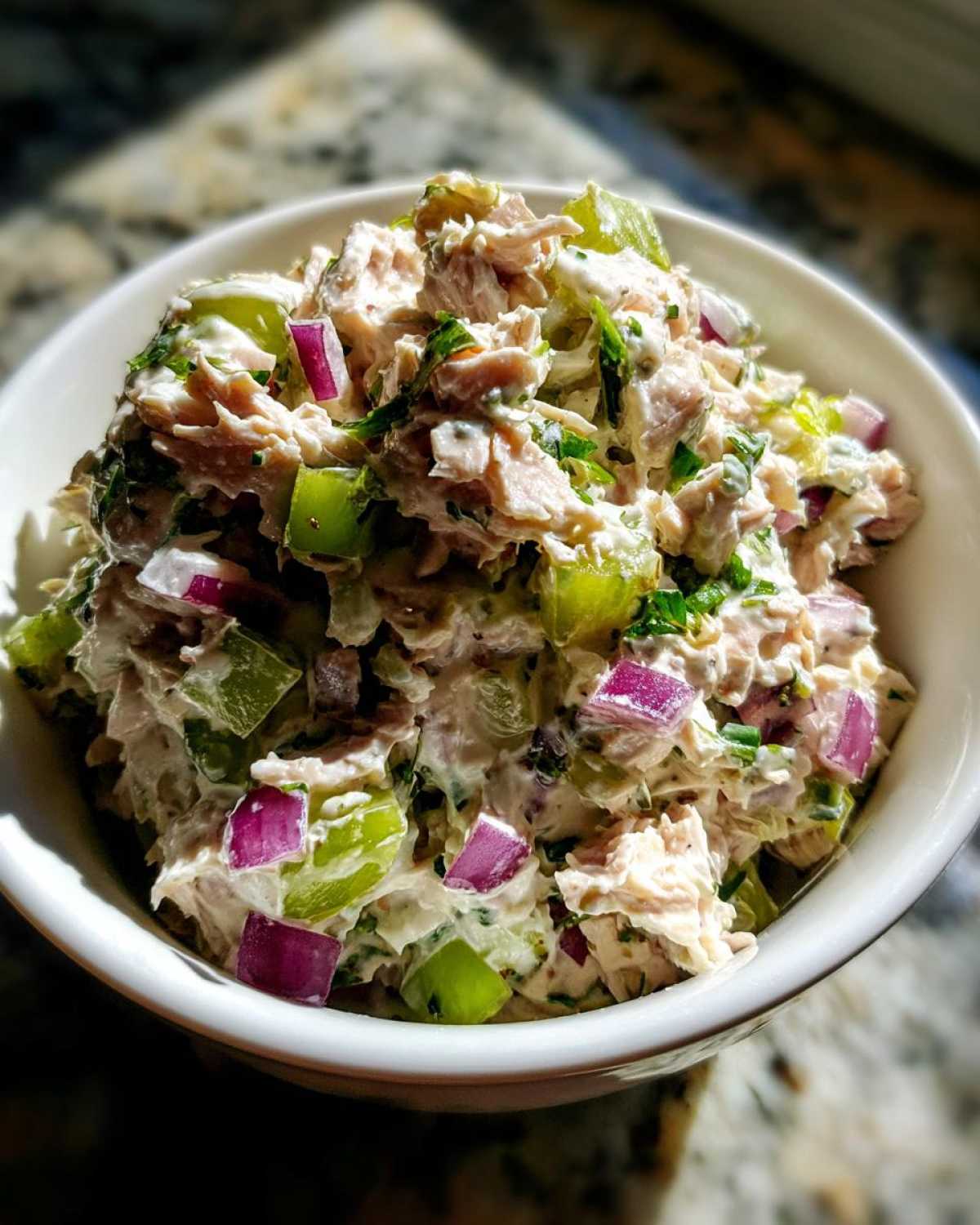 A close-up, brightly lit bowl filled with healthy tuna salad featuring chunks of tuna, celery, and diced red onion.