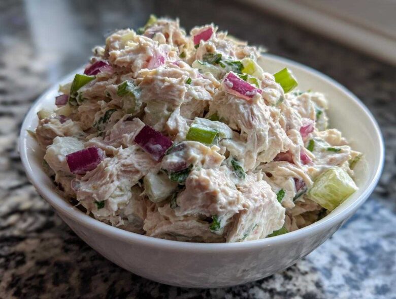 A close-up shot of creamy, chunky healthy tuna salad mixed with diced red onion and celery in a white bowl.
